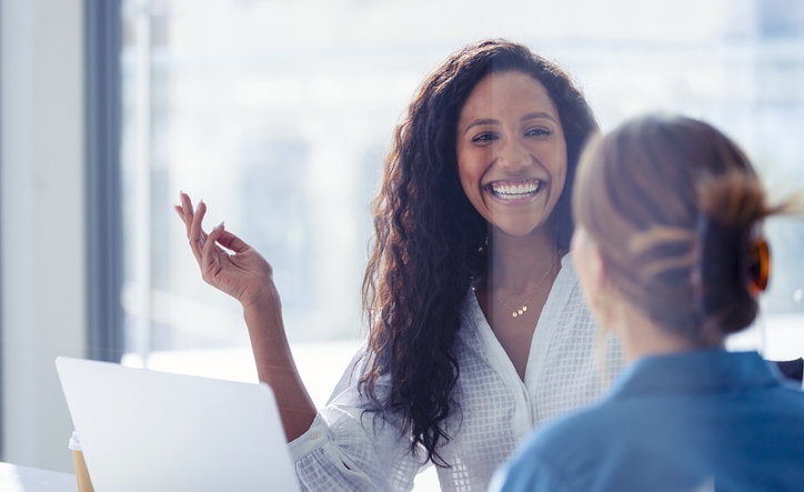 Business colleagues having a conversation. They are both young business people casually dressed in a modern office. Could be an interview or consultant working with a client. She is listening and smiling. One person has her back to us. Mixed ethnic group. One is African American and the other is Caucasian