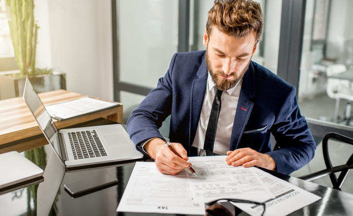 Handsome tax manager dressed in the suit working with documents and laptop at the modern office interior